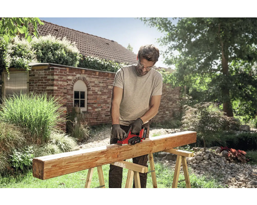 Ein Mann nutzt einen Elektrohobel an einem Holzbalken auf Holzböcken im hellen Garten. Er trägt eine Schutzbrille und Handschuhe.