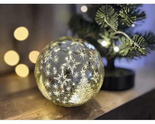 Boule en verre LED illuminée avec motifs d'étoiles et de sapin de Noël en aspect craquelé sur surface en bois à côté d'un petit sapin de Noël.