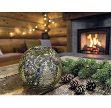 Boule LED décorative en verre avec sapins et étoiles sur une table en bois avec pommes de pin et branches devant une cheminée dans un chalet.