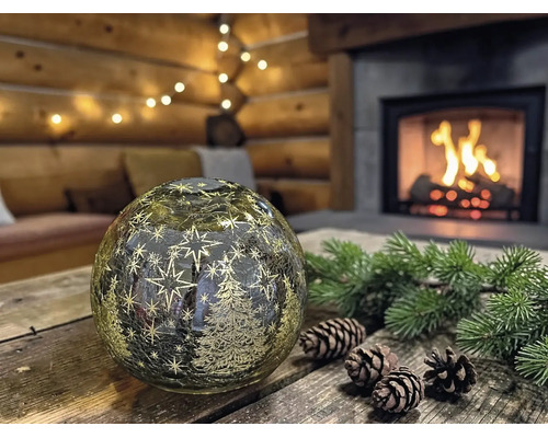 Boule LED décorative en verre avec sapins et étoiles sur une table en bois avec pommes de pin et branches devant une cheminée dans un chalet.