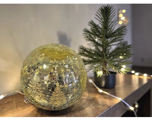 Boule en verre éclairée avec effet craquelé, étoiles et sapins sur une surface en bois à côté d'un sapin artificiel.