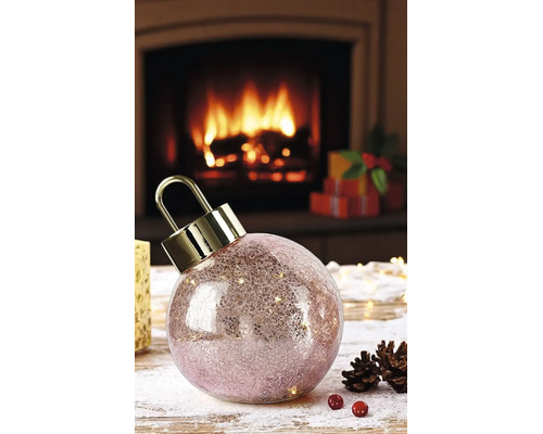 Boule de Noël à LED en verre craquelé avec attache dorée devant une cheminée allumée avec des pommes de pin et des cadeaux.