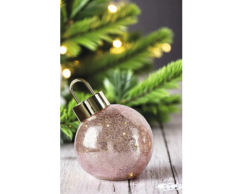 Boule de Noël en verre texturé rose avec un capuchon doré devant un sapin de Noël illuminé.