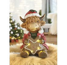 Figurine de Noël vache Highland avec bonnet de Noël et étoile lumineuse sur un tapis en fourrure devant un sapin de Noël décoré.