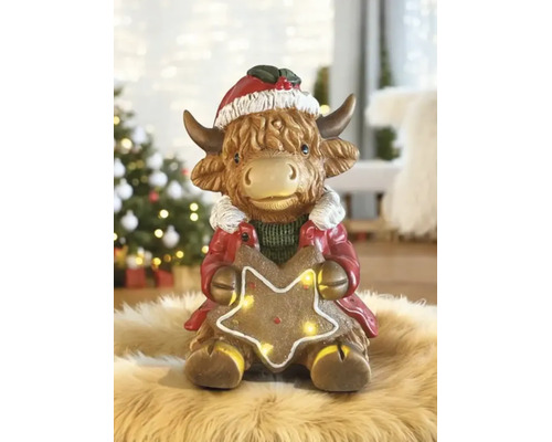 Figurine de Noël vache Highland avec bonnet de Noël et étoile lumineuse sur un tapis en fourrure devant un sapin de Noël décoré.