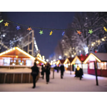 Lichterkette mit bunten Sternen vor einem beleuchteten Weihnachtsmarkt mit verschneiten Buden bei Nacht.