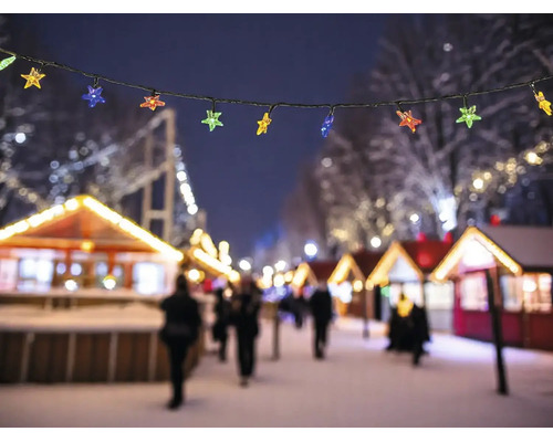 Lichterkette mit bunten Sternen vor einem beleuchteten Weihnachtsmarkt mit verschneiten Buden bei Nacht.