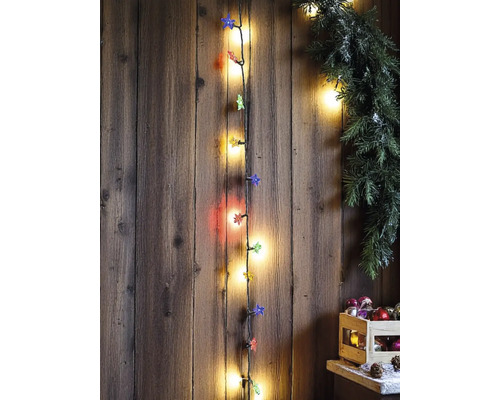 Vertikale Lichterkette mit Sternenaufsätzen an einer rustikalen Holzwand, dekoriert mit einer grünen Girlande und Weihnachtsschmuck in einer Kiste.