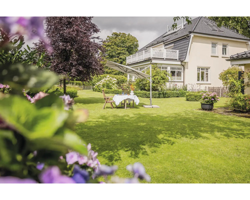 Un grand jardin avec pelouse, mobilier de jardin en bois et un grand parasol déporté gris devant une maison.