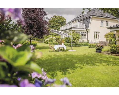 Großer weißer Ampelschirm über einem Gartentisch mit Stühlen auf einem hellen, weitläufigen Rasen vor einem Wohnhaus.
