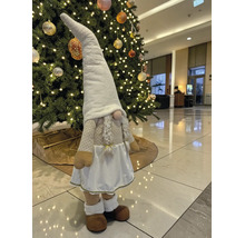 Dekorative Weihnachts-Wichtelfigur aus Stoff mit langer Mütze und Zöpfen in einem hellen Raum vor einem beleuchteten Weihnachtsbaum.