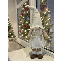 Wichtel-Figur mit spitzem Hut, Zöpfen und Rock auf einer Fensterbank vor einem hellen, festlich geschmückten Weihnachtsbaum.