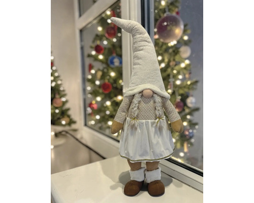 Wichtel-Figur mit spitzem Hut, Zöpfen und Rock auf einer Fensterbank vor einem hellen, festlich geschmückten Weihnachtsbaum.