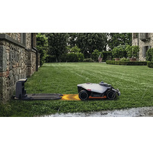 Robot tondeuse Mammotion sur une pelouse verte approchant de la station de charge sous la pluie devant un mur en pierre.