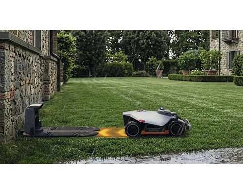 Robot tondeuse Mammotion sur une pelouse verte approchant de la station de charge sous la pluie devant un mur en pierre.