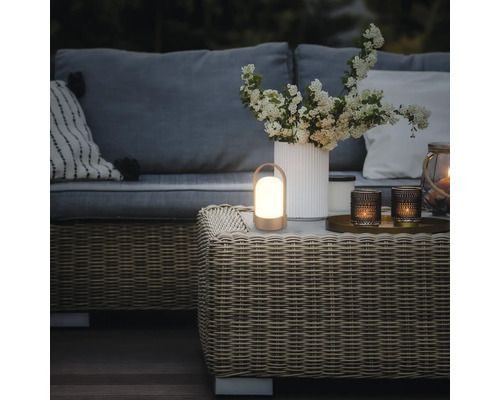 Lanterne LED portable sur une table basse en résine tressée dans un salon de jardin avec canapé, coussins et vase de fleurs.