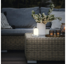 Lampe de table LED portable à lumière chaude sur une table en rotin dans un salon de jardin moderne avec canapé et fleurs.