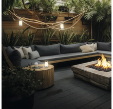 Salon de jardin moderne avec canapé gris, coussins, foyer, table d appoint en bois avec lanterne et éclairage d ambiance en soirée.