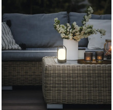 Lampe de table à LED portable sur une table en rotin dans un salon de jardin chaleureusement éclairé avec canapé, coussins et vase de fleurs.