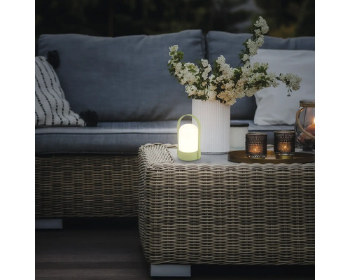 Lampe de table LED portable sur une table de jardin tressée dans un espace extérieur chaleureux avec canapé et fleurs.