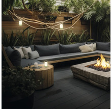 Espace extérieur moderne avec canapé en L, foyer en bois et lanternes sur une terrasse sombre, entouré de plantes et une clôture en bois.