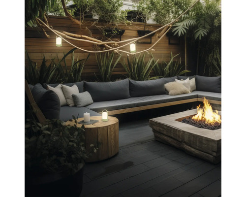 Espace extérieur moderne avec canapé en L, foyer en bois et lanternes sur une terrasse sombre, entouré de plantes et une clôture en bois.
