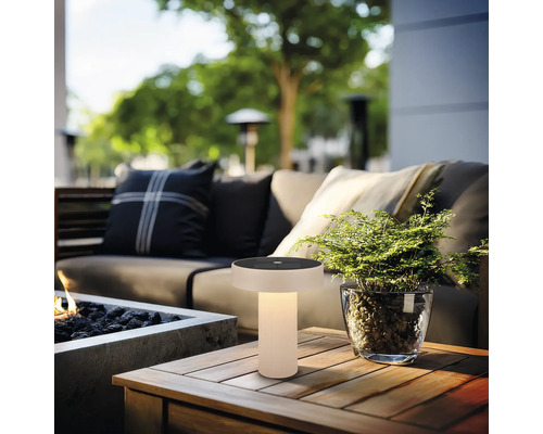 Lampe de table solaire d’extérieur moderne avec lumière chaude sur une table en bois dans un salon de jardin avec canapé et plante.
