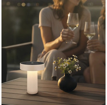 Lampe de table solaire moderne sur une table de terrasse en bois dans un jardin au crépuscule avec des fleurs et des personnes en arrière-plan.