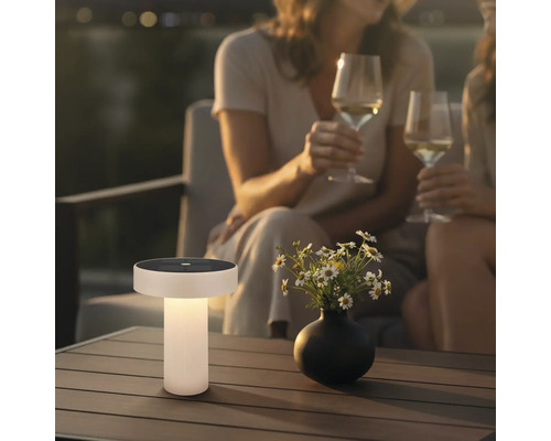 Lampe de table solaire moderne sur une table de terrasse en bois dans un jardin au crépuscule avec des fleurs et des personnes en arrière-plan.