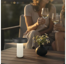 Lampe de table solaire moderne sur une table de terrasse à côté d'un vase de fleurs, en arrière-plan deux personnes avec des verres de vin.