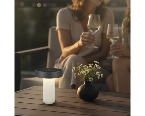 Lampe de table solaire moderne sur une table de terrasse à côté d'un vase de fleurs, en arrière-plan deux personnes avec des verres de vin.