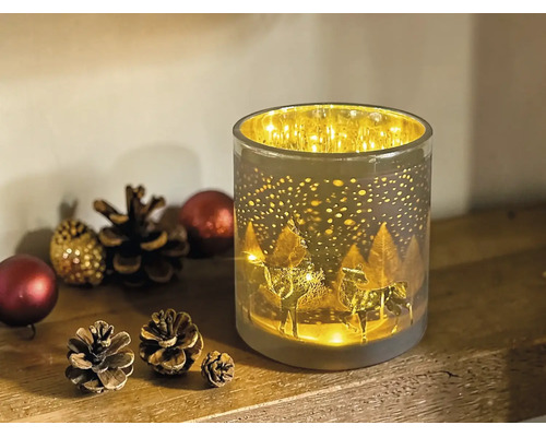 Zylindrisches Windlicht aus Glas mit beleuchtetem Rentier- und Waldmotiv, dekoriert mit Tannenzapfen auf einer Holzoberfläche.