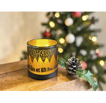 Dekoratives Windlicht aus Glas mit goldenem Waldmotiv auf einer Holzoberfläche, im Hintergrund ein geschmückter Weihnachtsbaum.
