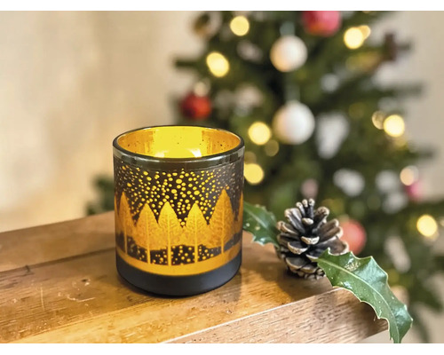 Dekoratives Windlicht aus Glas mit goldenem Waldmotiv auf einer Holzoberfläche, im Hintergrund ein geschmückter Weihnachtsbaum.