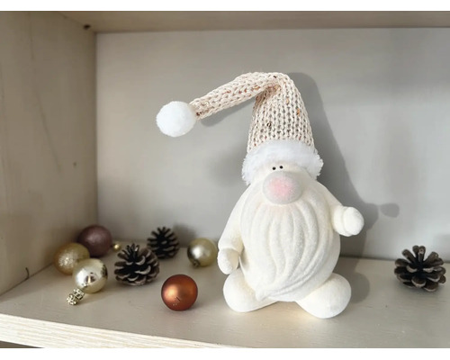 Dekorativer weißer Weihnachtswichtel mit Strickmütze und weißem Bart auf einem Regal mit Weihnachtskugeln und Tannenzapfen.