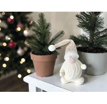 Weihnachts-Wichtel mit Strickmütze neben kleinen Tannen in Töpfen auf weißem Tisch vor geschmücktem Weihnachtsbaum.