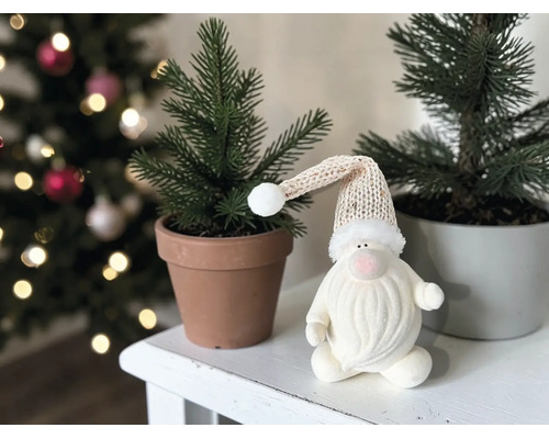 Weihnachts-Wichtel mit Strickmütze neben kleinen Tannen in Töpfen auf weißem Tisch vor geschmücktem Weihnachtsbaum.