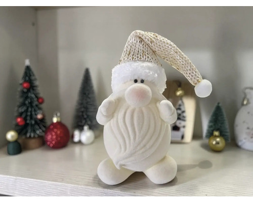 Weiße Plüschfigur eines Weihnachtsmanns mit Bart und langer Strickmütze auf einem Regal neben kleinen Tannenbäumen und Weihnachtsschmuck.