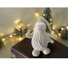 Dekorative Weihnachtsmannfigur mit Daumen-hoch-Geste auf einem Koffer vor einem beleuchteten Weihnachtsbaum.