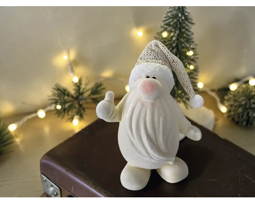 Dekorative Weihnachtsmannfigur mit Daumen-hoch-Geste auf einem Koffer vor einem beleuchteten Weihnachtsbaum.