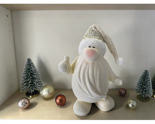 Kleine weiße Weihnachtsmann-Figur mit Daumen nach oben, Strickmütze, kleinen Tannenbäumen und Weihnachtskugeln auf einer Ablage.