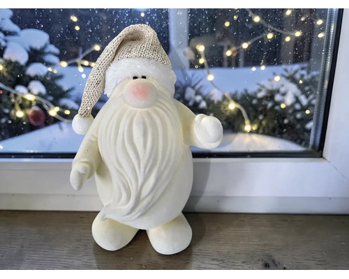 Weihnachtsmann-Dekofigur mit langem Bart und glitzernder Mütze auf Fensterbank vor verschneitem Hintergrund mit Lichterkette.