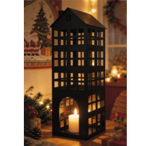 Lanterne en métal noir en forme de maison avec bougie blanche allumée dans un décor de Noël festif avec branches de sapin et lumières chaudes.