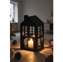 Lanterne en forme de maison en métal noir avec une bougie allumée, entourée de décorations de Noël et de pommes de pin dans une pièce lumineuse.