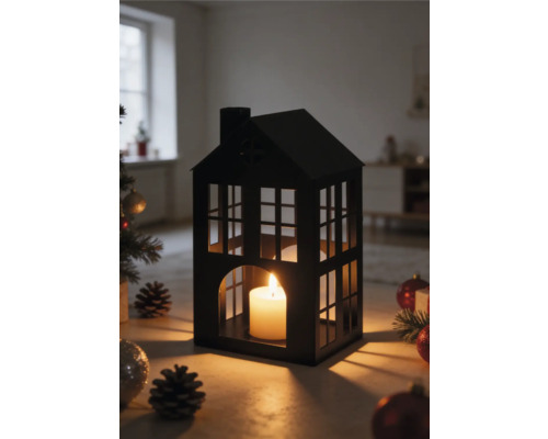 Lanterne en forme de maison en métal noir avec une bougie allumée, entourée de décorations de Noël et de pommes de pin dans une pièce lumineuse.