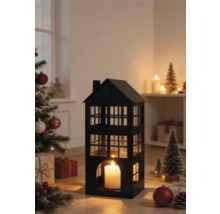 Lanterne noire en métal en forme de maison avec une bougie allumée dans une scène de Noël avec un sapin décoré et des cadeaux.