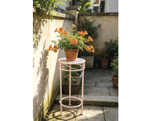 Hoher Blumenhocker aus Metall mit einer blühenden Klettertrompete im Tontopf auf einer hellen Terrasse.