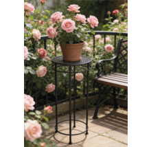 Hoher runder Blumenständer aus schwarzem Metall mit Ornamenten darauf ein Tontopf mit rosa Rosen in einem hellen Garten neben einer Sitzbank.