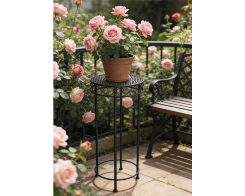 Hoher runder Blumenständer aus schwarzem Metall mit Ornamenten darauf ein Tontopf mit rosa Rosen in einem hellen Garten neben einer Sitzbank.