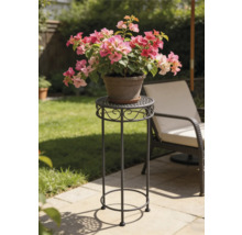 Hoher dekorativer Blumenständer aus Metall auf einer sonnigen Gartenterrasse mit runder perforierter Ablage und Schnörkelverzierung.